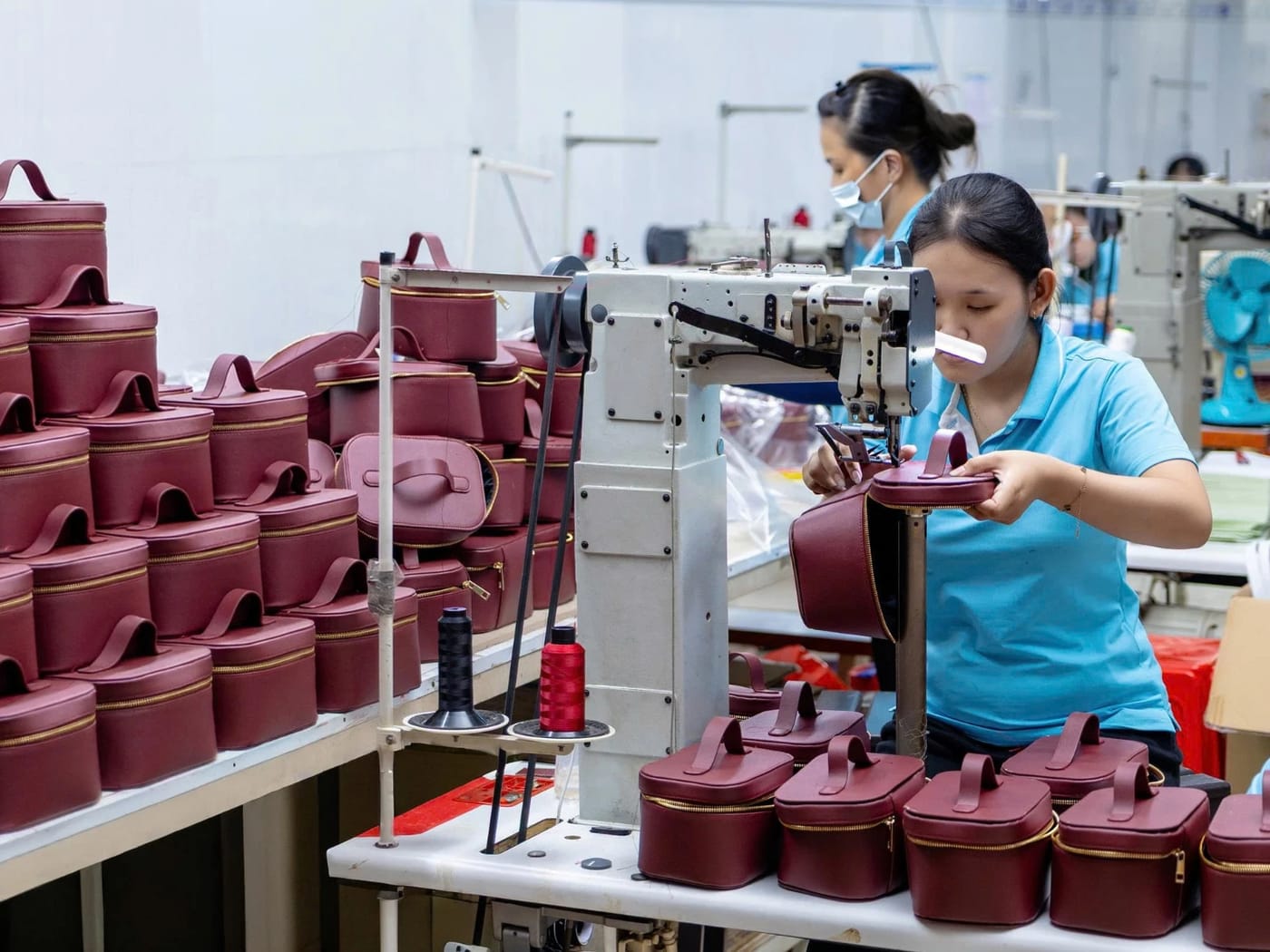 Seamstress stitching a burgundy cosmetic bag on an industrial sewing machine, with stacks of finished bags beside her, in the Hoang Nhat workshop